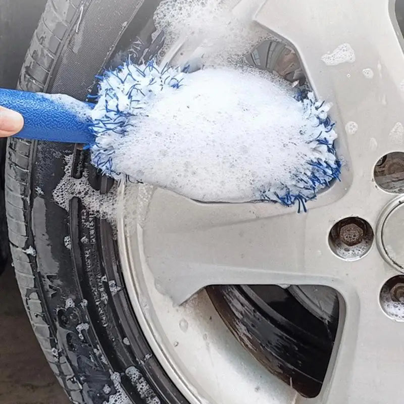 Cleaning a silver car wheel with a blue handled, soapy wash brush.